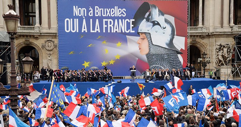 Supporters of France's far-right National Front (FN) wave French flags as leader Marine Le Pen (rear C) delivers a speech in front of a poster depicting Joan of Arc (Jeanne d'Arc) and reading "No to Brussels, yes to France" during a May Day rally in Paris on May 1, 2014. AFP PHOTO / PIERRE ANDRIEU        (Photo credit should read PIERRE ANDRIEU/AFP/Getty Images)